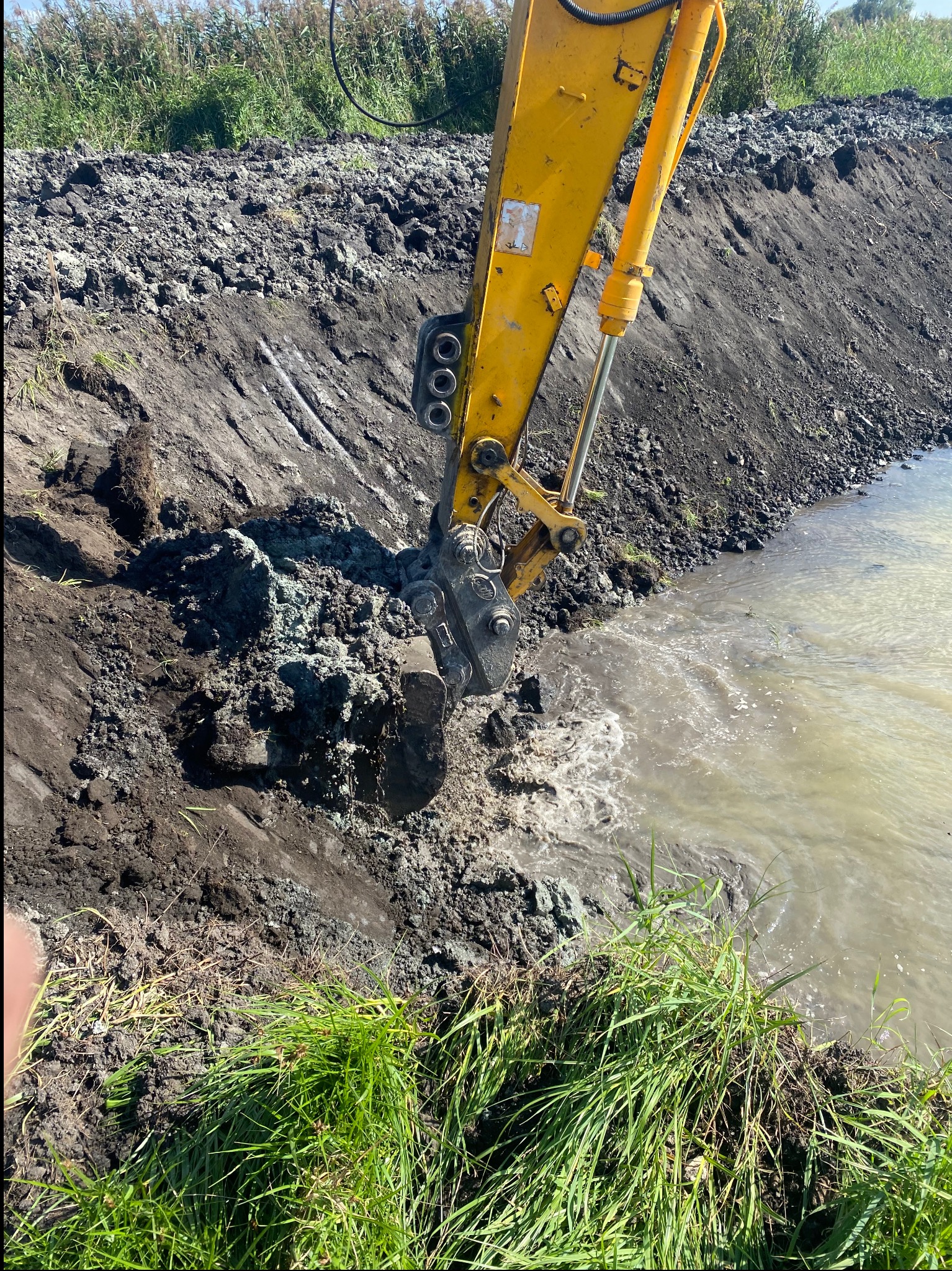 Żółta koparka pogłębia brzeg zbiornika wodnego, nabierając urobek. W tle widać skarpę z ziemi i roślinność. Prace ziemne w toku, z bliska.