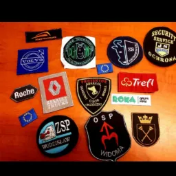 Kolekcja naszywek i emblematów różnych firm i organizacji, w tym Volvo, Roche, Renault Trucks, Trefl, Roka, a także emblematy szkół, agencji ochrony i OSP, ułożonych na drewnianym stole.