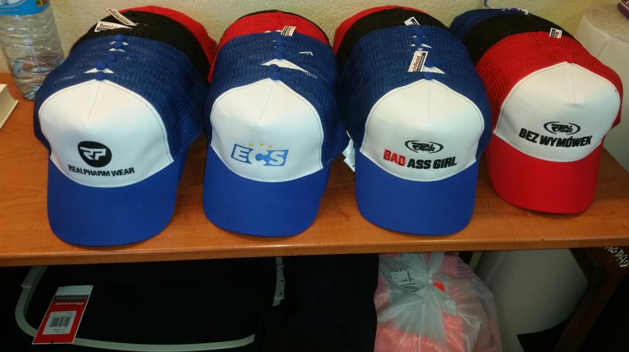 Rząd czapek z daszkiem, każda z unikalnym haftowanym logo: 'REALPHARM WEAR', 'ECS', 'BAD ASS GIRL', 'BEZ WYMÓWEK', w kolorach niebieskim, czerwonym i czarnym z białymi panelami przednimi.