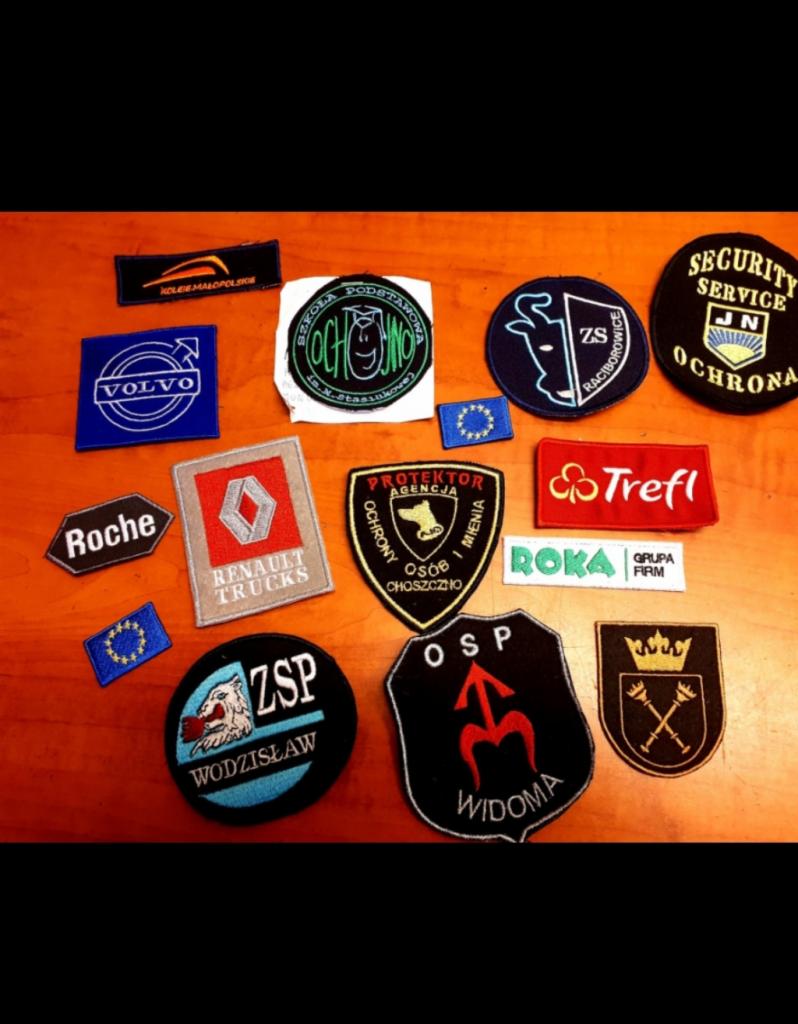 Kolekcja naszywek i emblematów różnych firm i organizacji, w tym Volvo, Roche, Renault Trucks, Trefl, Roka, a także emblematy szkół, agencji ochrony i OSP, ułożonych na drewnianym stole.