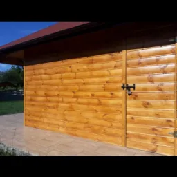 Drewniana elewacja budynku gospodarczego z brązowym dachem i czarnym zamkiem na drzwiach, widoczna z boku na tle zieleni.