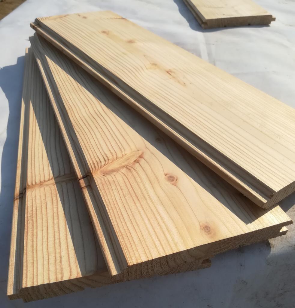 Stos drewnianych desek elewacyjnych z widocznymi sękami i frezowanymi krawędziami, ułożonych na białej folii budowlanej.