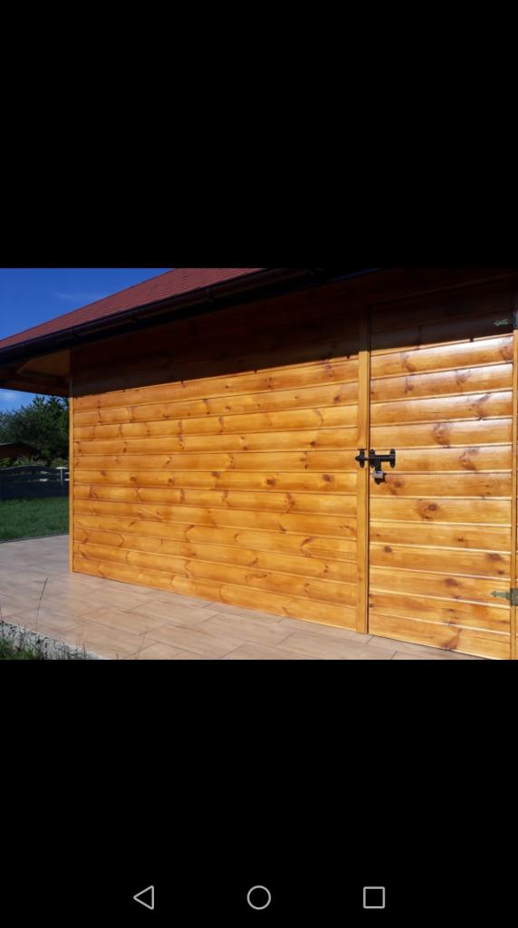 Drewniana elewacja budynku gospodarczego z brązowym dachem i czarnym zamkiem na drzwiach, widoczna z boku na tle zieleni.