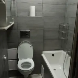 Szara łazienka z białą armaturą: umywalka, toaleta z otwartą klapą, wanna z baterią, szafka z lustrem, półka na kosmetyki i rolka papieru toaletowego.