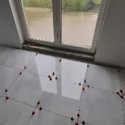 Układanie jasnoszarych płytek podłogowych z użyciem systemu poziomowania, widok na okno z deszczową pogodą na zewnątrz.