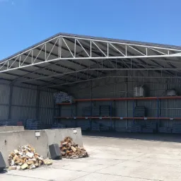 Otwarta hala magazynowa z konstrukcją stalową, wypełniona materiałami budowlanymi i opałowymi, w tle błękitne niebo.