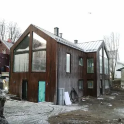 Nowoczesny dom z ciemnego drewna z dużymi oknami, w trakcie budowy, z widocznymi elementami konstrukcyjnymi i zabezpieczoną folią turkusową ościeżnicą drzwiową, na tle sąsiednich budynków i nieba...