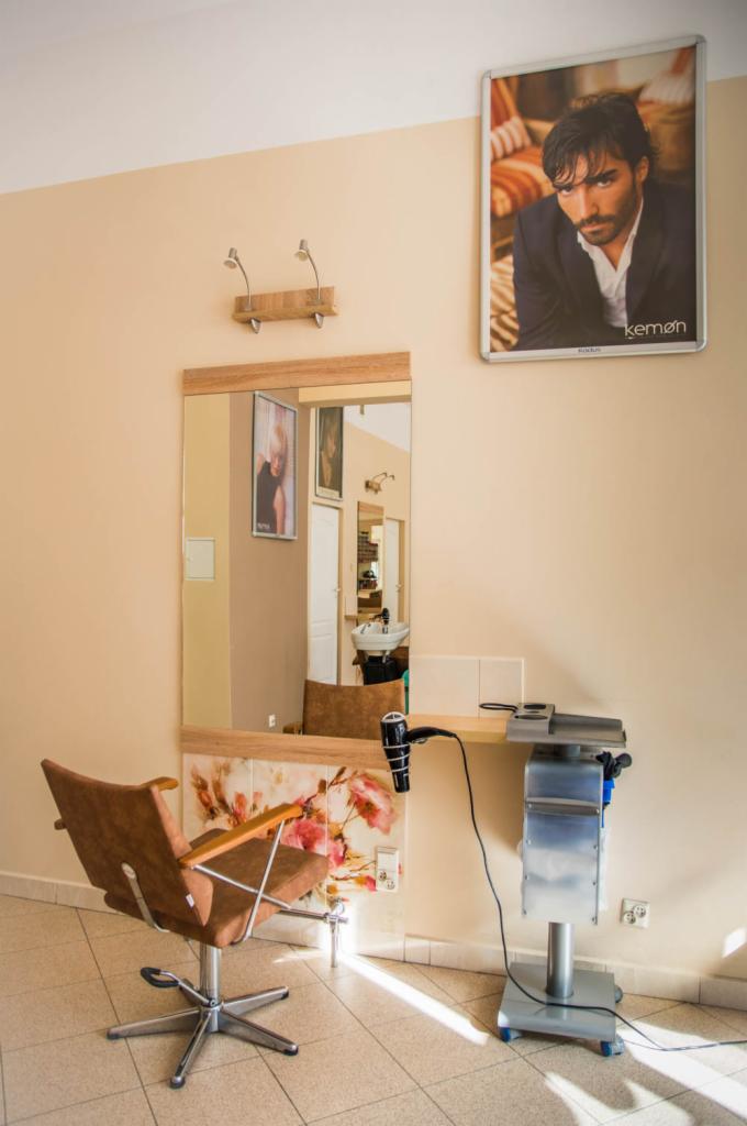 Stanowisko z lustrem i fotelem w salonie, na ścianie plakat reklamowy z wizerunkiem mężczyzny, suszarka do włosów na blacie.