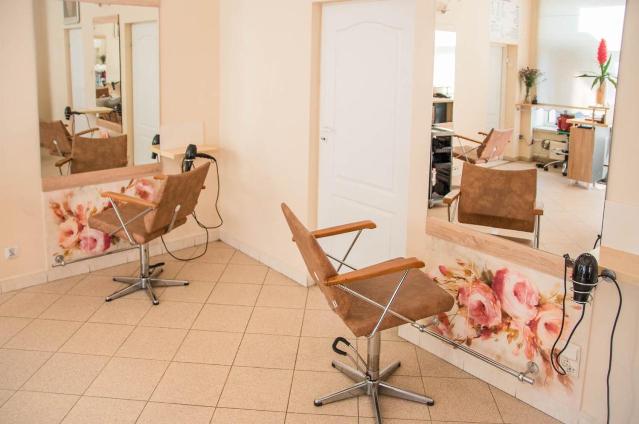 Dwa puste stanowiska z brązowymi fotelami obrotowymi i suszarkami w salonie, lustra z motywem kwiatowym, beżowe płytki na podłodze.