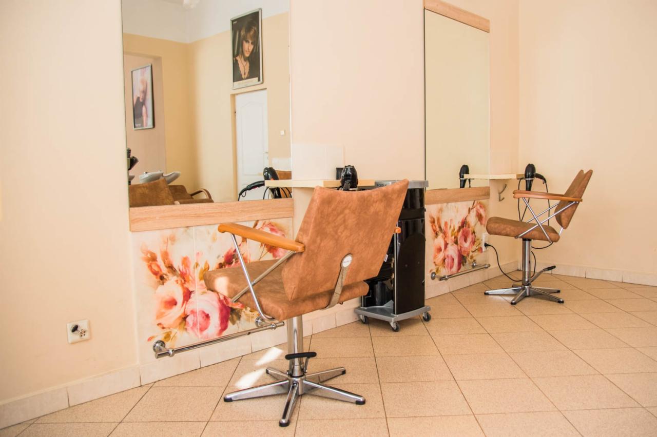 Puste stanowisko z brązowym fotelem fryzjerskim i lustrem w salonie, ściana zdobiona motywem kwiatowym, w tle widoczna myjnia fryzjerska.