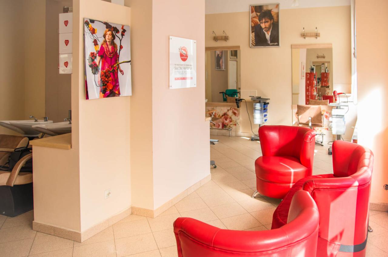 Jasne wnętrze salonu z czerwonymi fotelami, umywalkami fryzjerskimi i plakatami na ścianach.