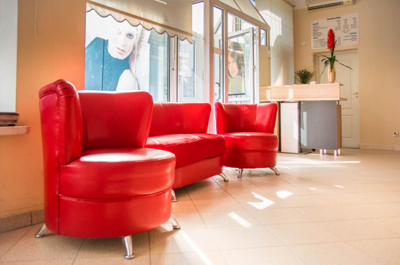 Poczekalnia salonu fryzjerskiego z czerwonymi, skórzanymi fotelami i kanapą, plakaty z wizerunkami kobiet w oknach, recepcja z kwiatami w tle.