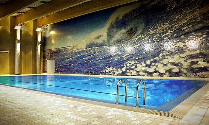 Kryty basen z turkusową wodą, metalowe drabinki wejściowe, ściana z mozaiką przedstawiającą falę i surfera, oświetlenie sufitowe, beżowe płytki podłogowe.