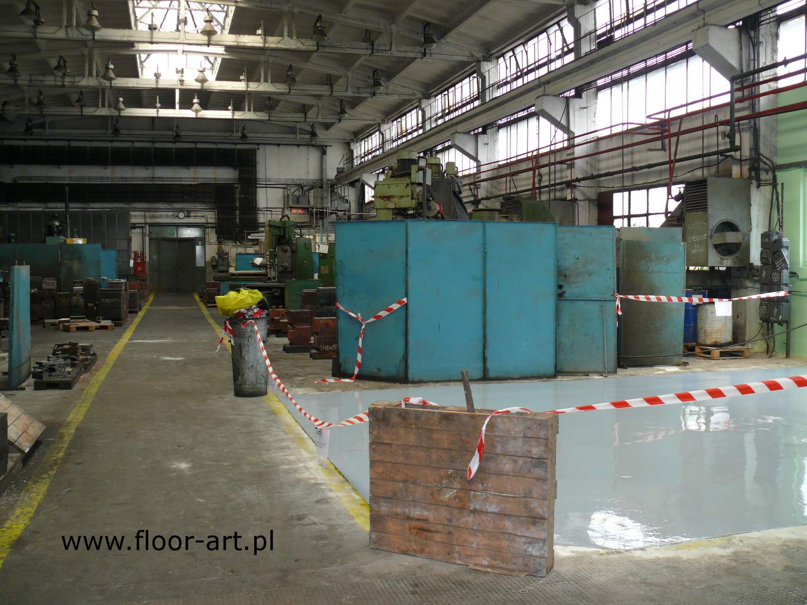 Szara posadzka żywiczna w hali produkcyjnej, odgrodzona taśmą ostrzegawczą, z widocznymi maszynami i elementami wyposażenia przemysłowego.