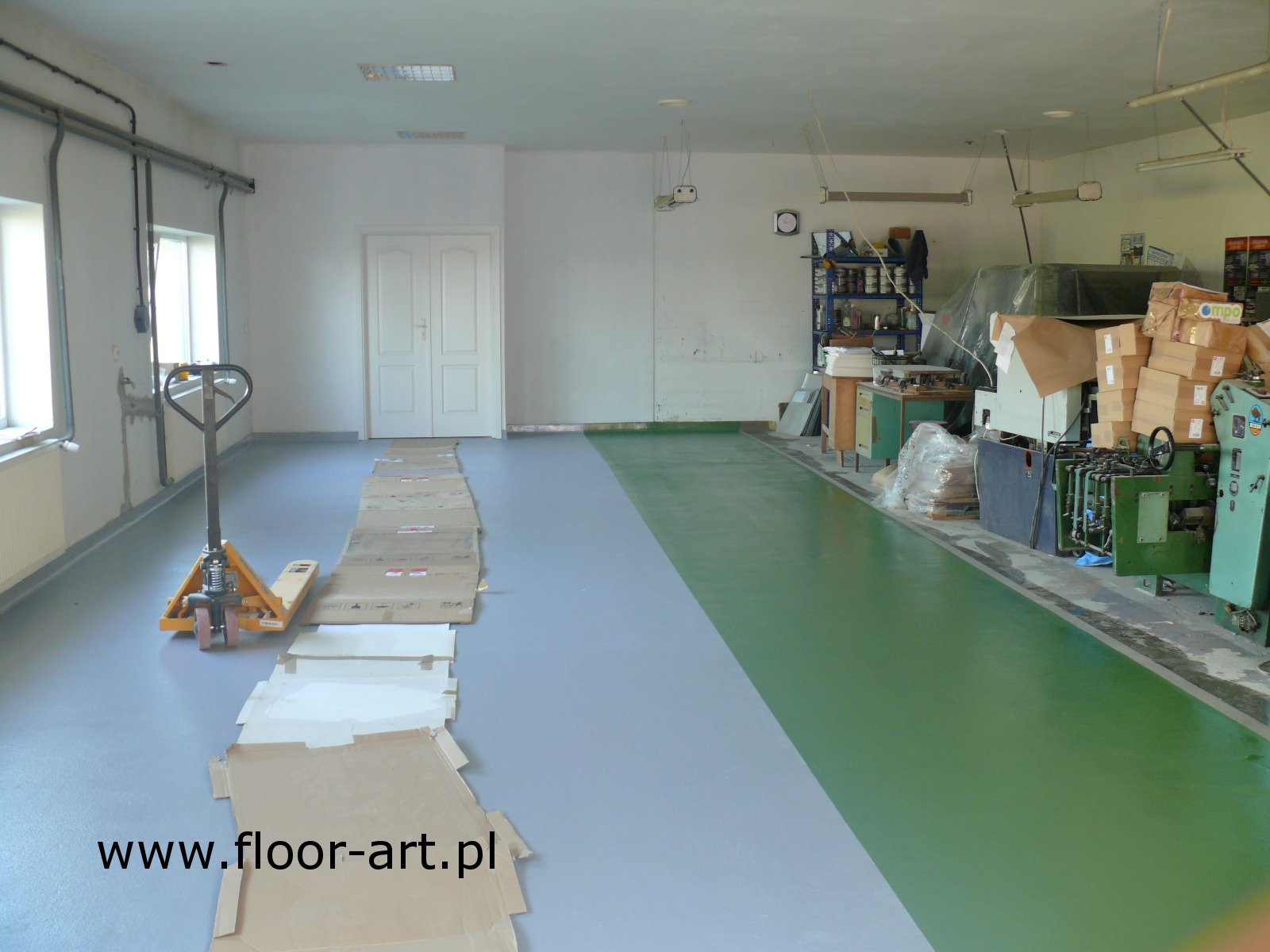 Szara posadzka żywiczna w hali produkcyjnej, częściowo zabezpieczona kartonami, z wózkiem paletowym obok okna, widoczna zielona strefa podłogi.