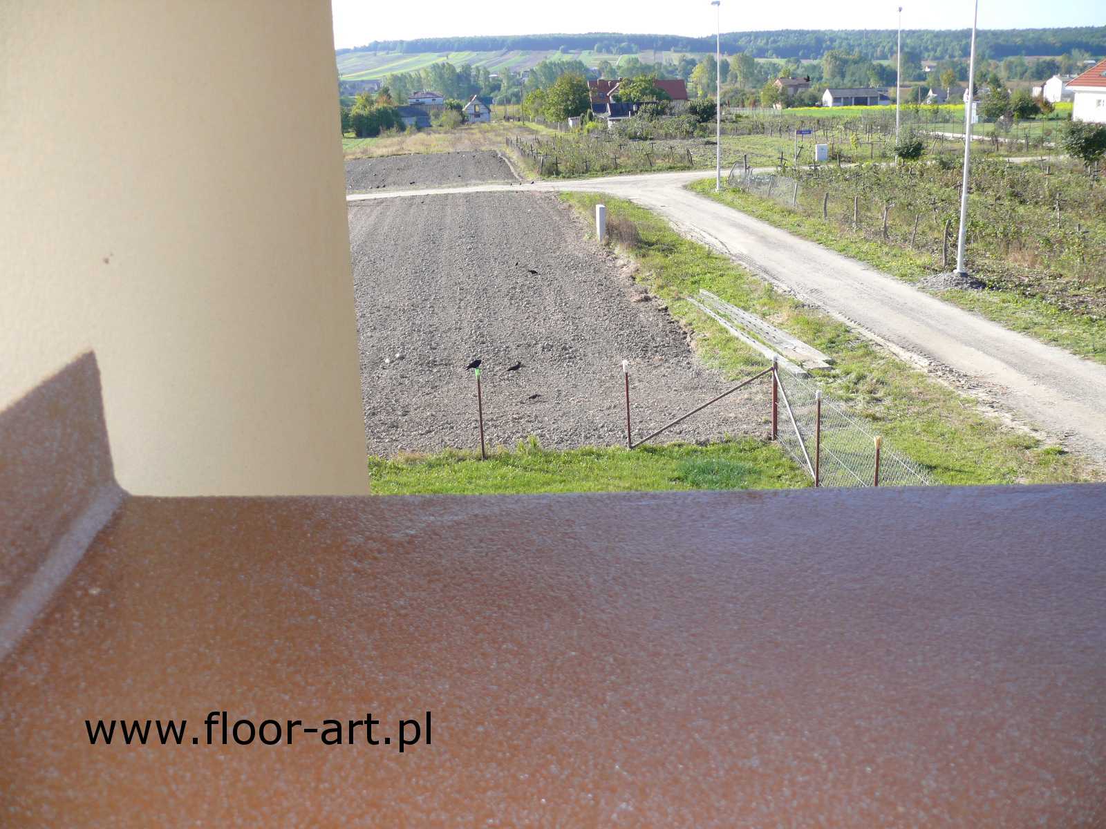 Balkon z żywicznej posadzki poliuretanowej w kolorze brązowym, widok na wiejski krajobraz z polami i drogą.