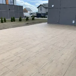 Ułożona kostka brukowa imitująca drewno na tarasie domu, widoczne tuje i budynki w tle.