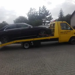 Czarny samoch&oacute;d osobowy zabezpieczony pasami transportowymi na ż&oacute;łtej platformie pojazdu pomocy drogowej z napisem 'NORBI AUTOHOL' i numerem telefonu, na tle brukowanej nawierzchni i zabudowań.