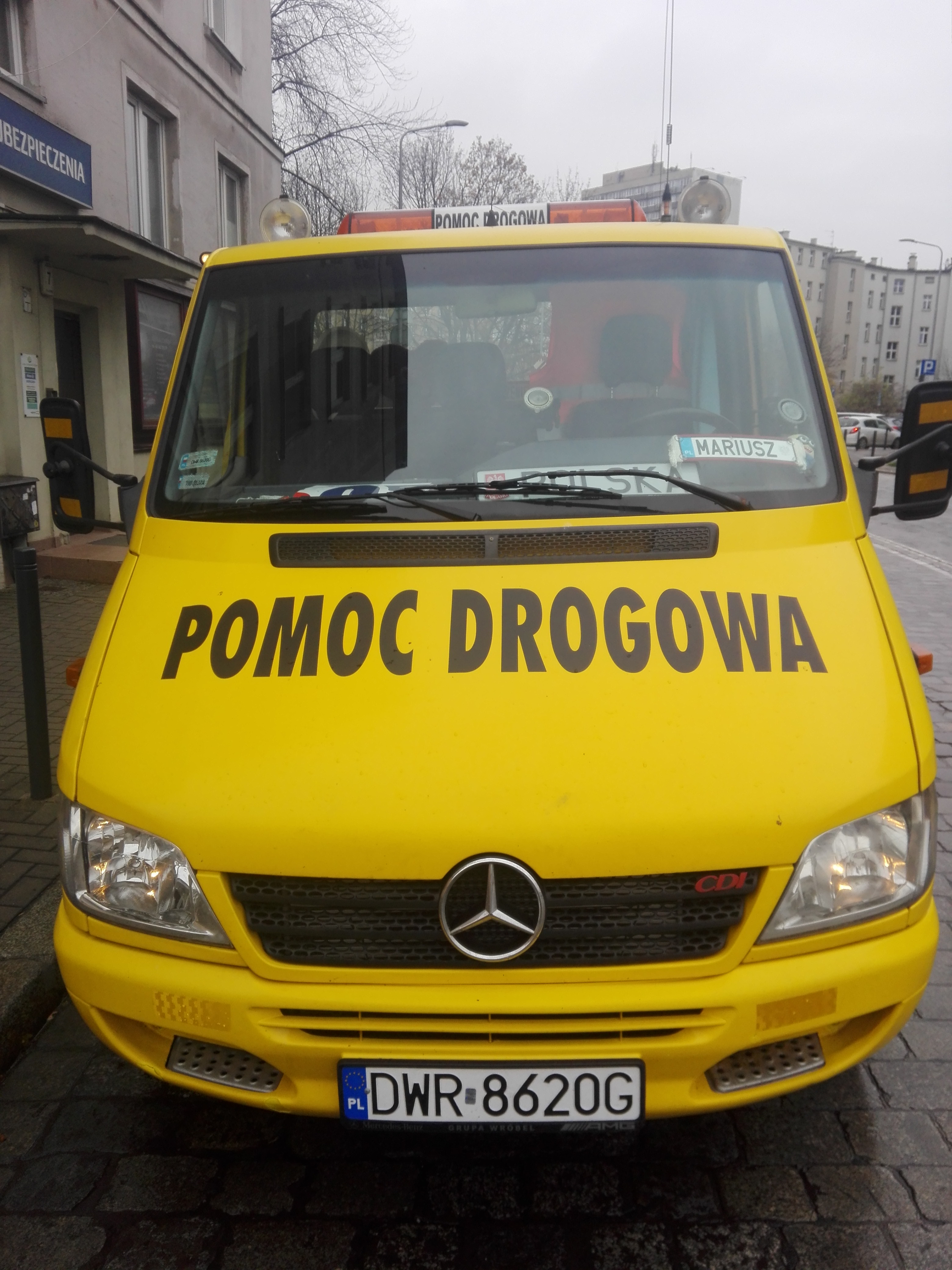 Żółty Mercedes Sprinter z napisem 'POMOC DROGOWA' na masce i tablicą rejestracyjną DWR 8620G, zaparkowany na ulicy w pochmurny dzień.
