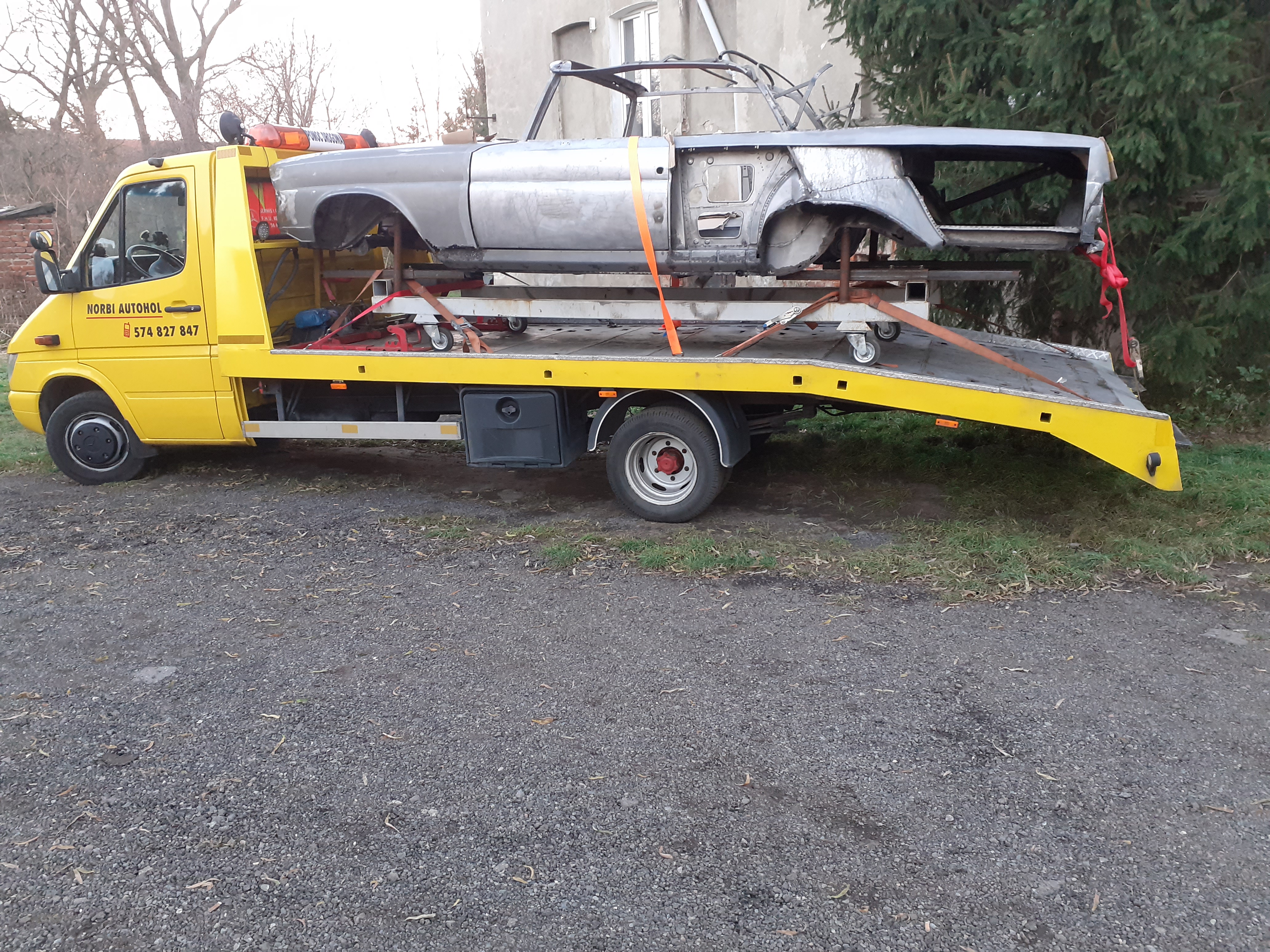 Żółty pojazd transportowy z nadwoziem samochodu osobowego bez karoserii, zabezpieczonym pomarańczowymi pasami, zaparkowany na żwirowym podłożu w pobliżu budynku i drzew.