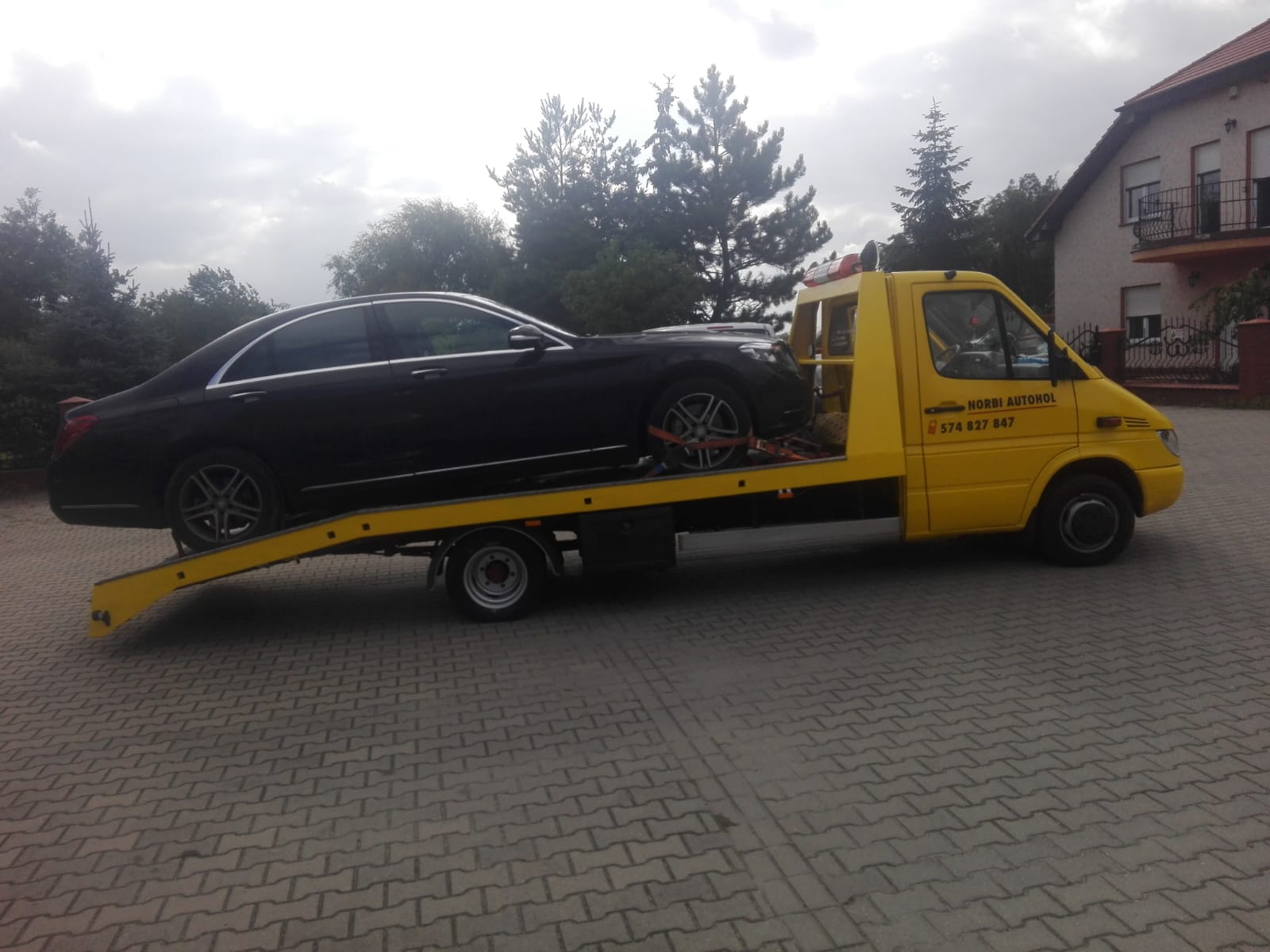 Czarny samochód osobowy zabezpieczony pasami transportowymi na żółtej platformie pojazdu pomocy drogowej z napisem 'NORBI AUTOHOL' i numerem telefonu, na tle brukowanej nawierzchni i zabudowań.