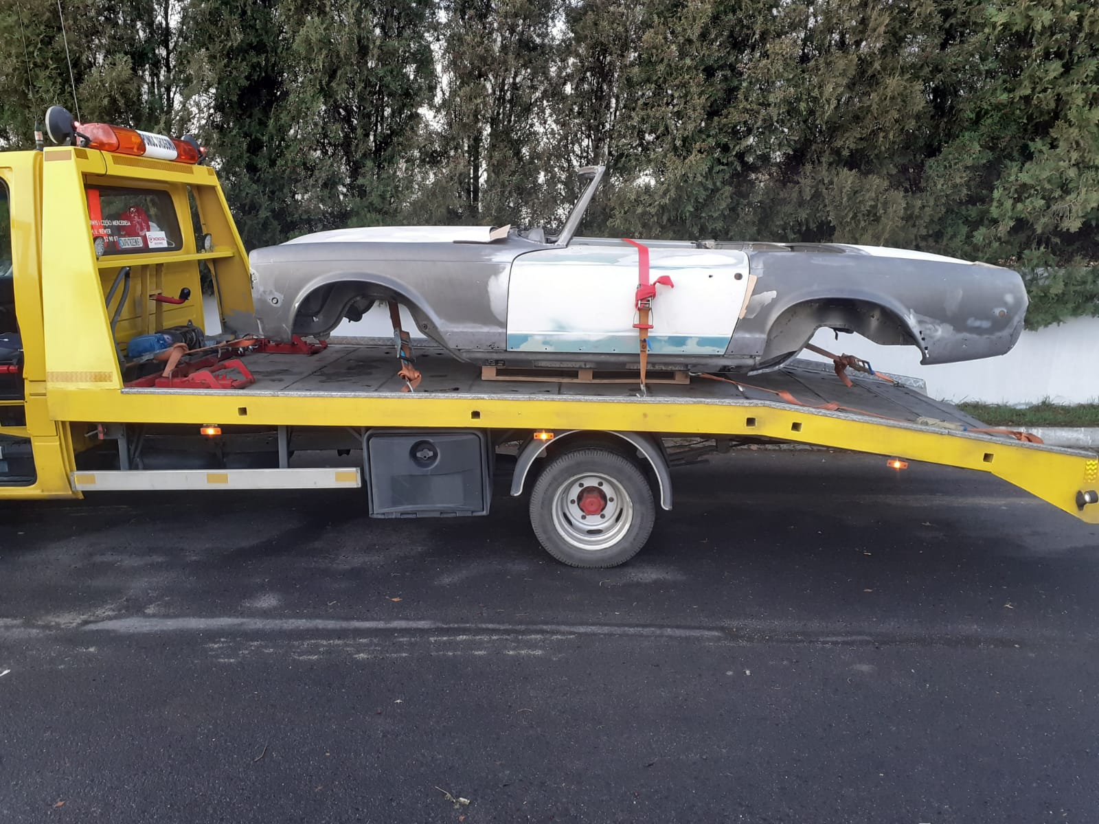 Żółta platforma transportowa przewozi karoserię zabytkowego samochodu cabrio w trakcie renowacji, zabezpieczoną czerwonymi pasami.