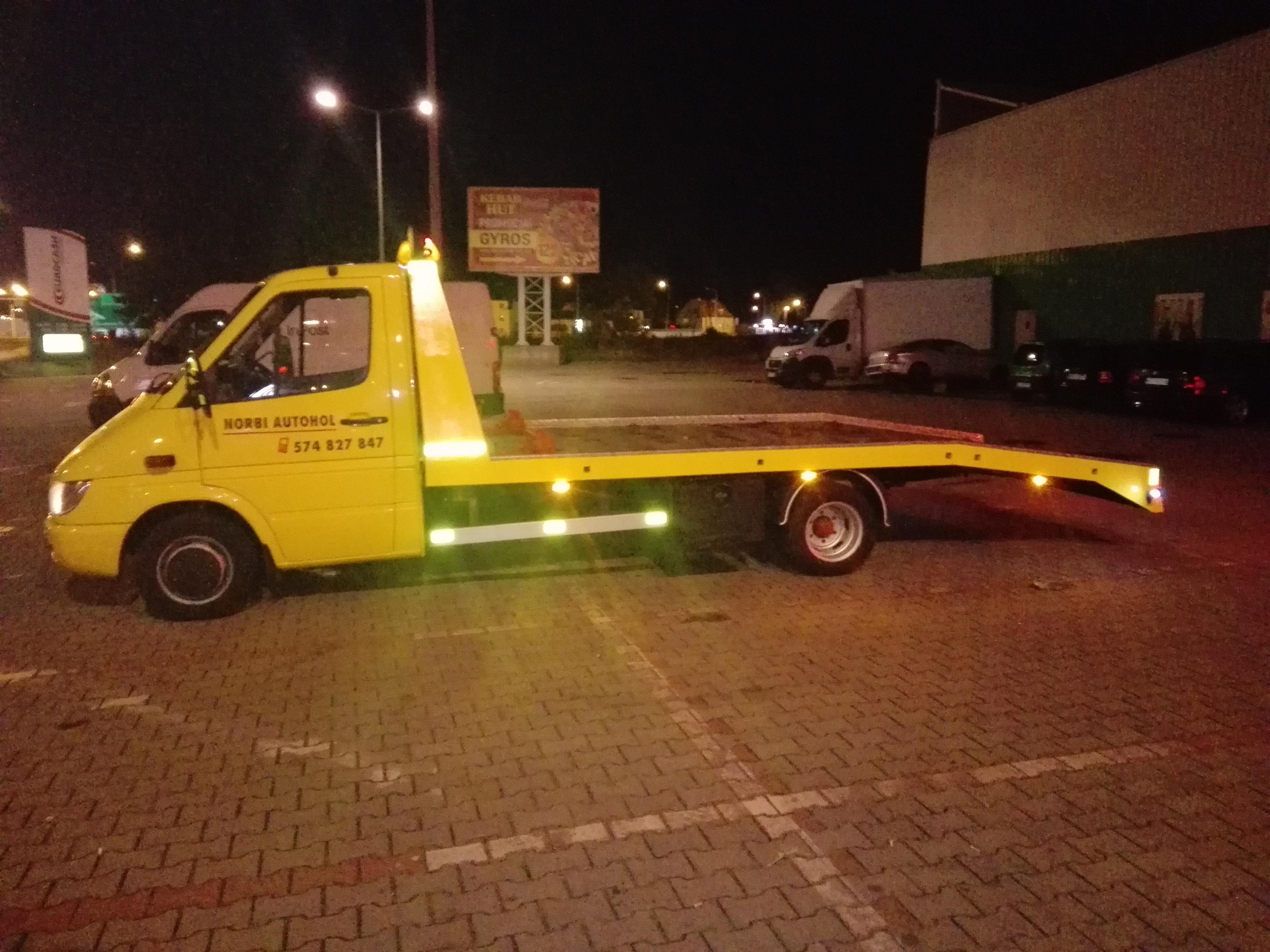 Żółty pojazd pomocy drogowej marki Mercedes-Benz Sprinter z numerem telefonu, zaparkowany na kostce brukowej w nocy, z reklamą gyrosa w tle.
