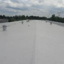 Jasnoszary, płaski dach przemysłowy z kominkami wentylacyjnymi, widoczna osoba wykonująca prace na dachu w oddali, w tle budynki i słupy wysokiego napięcia.