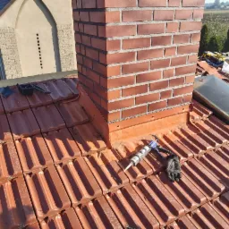 Ceglany komin na dachu z dachówką ceramiczną, obróbka blacharska wokół komina, narzędzia dekarskie i rękawica na dachu, okno dachowe w tle.