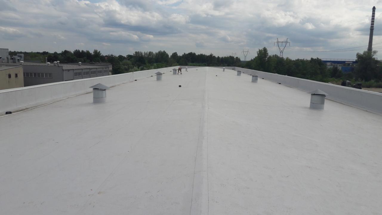 Jasnoszary, płaski dach przemysłowy z kominkami wentylacyjnymi, widoczna osoba wykonująca prace na dachu w oddali, w tle budynki i słupy wysokiego napięcia.