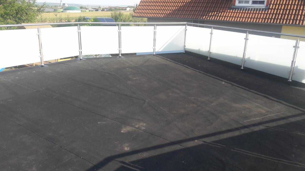 Wykończony dach płaski z czarną membraną, zabezpieczony balustradą ze stali nierdzewnej i mlecznego szkła, widok na okoliczne budynki i drzewa w tle.