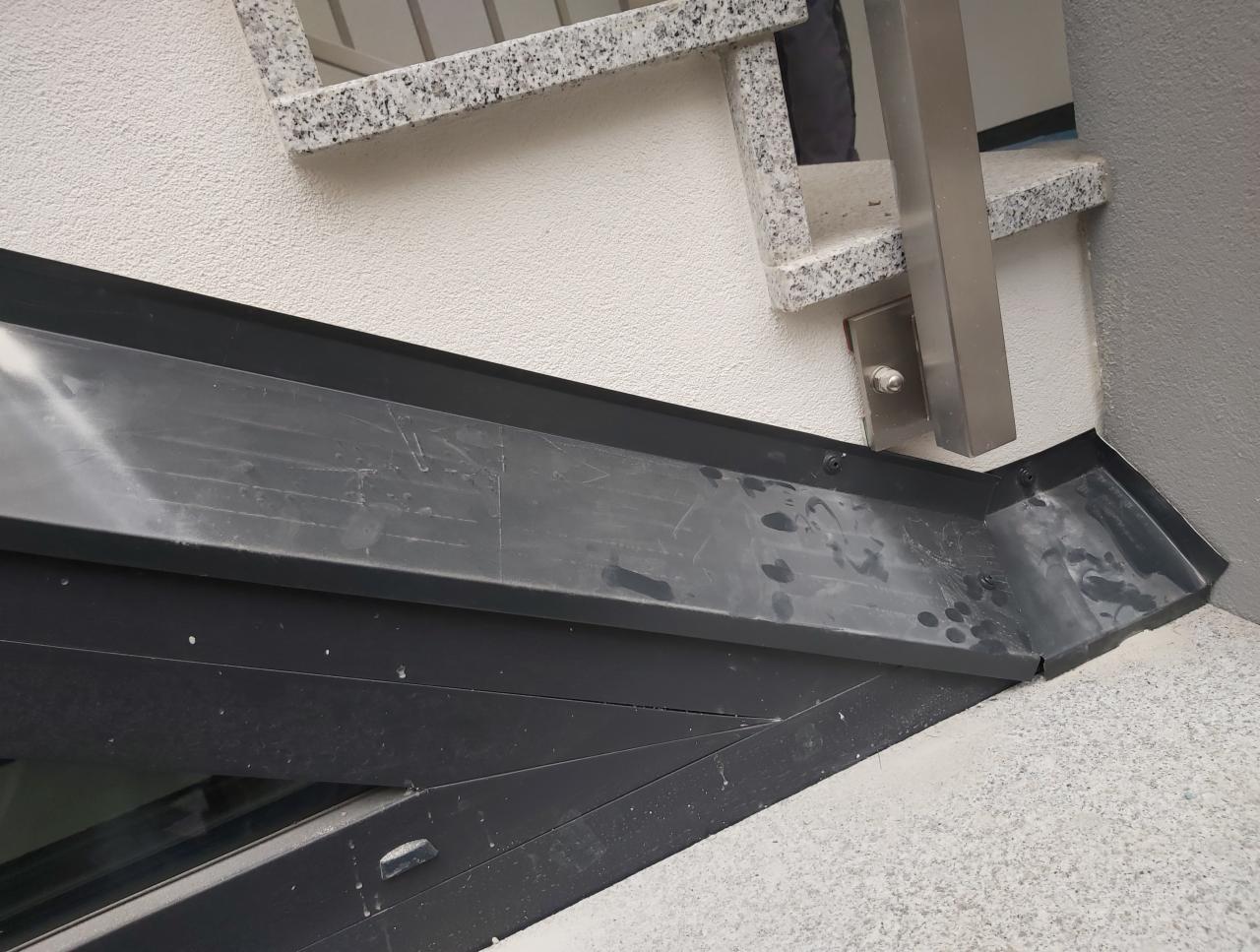 Szczegółowe ujęcie czarnej, metalowej rynny przy styku ze ścianą budynku i schodami z granitowym wykończeniem, widoczne ślady po wodzie i montażu.