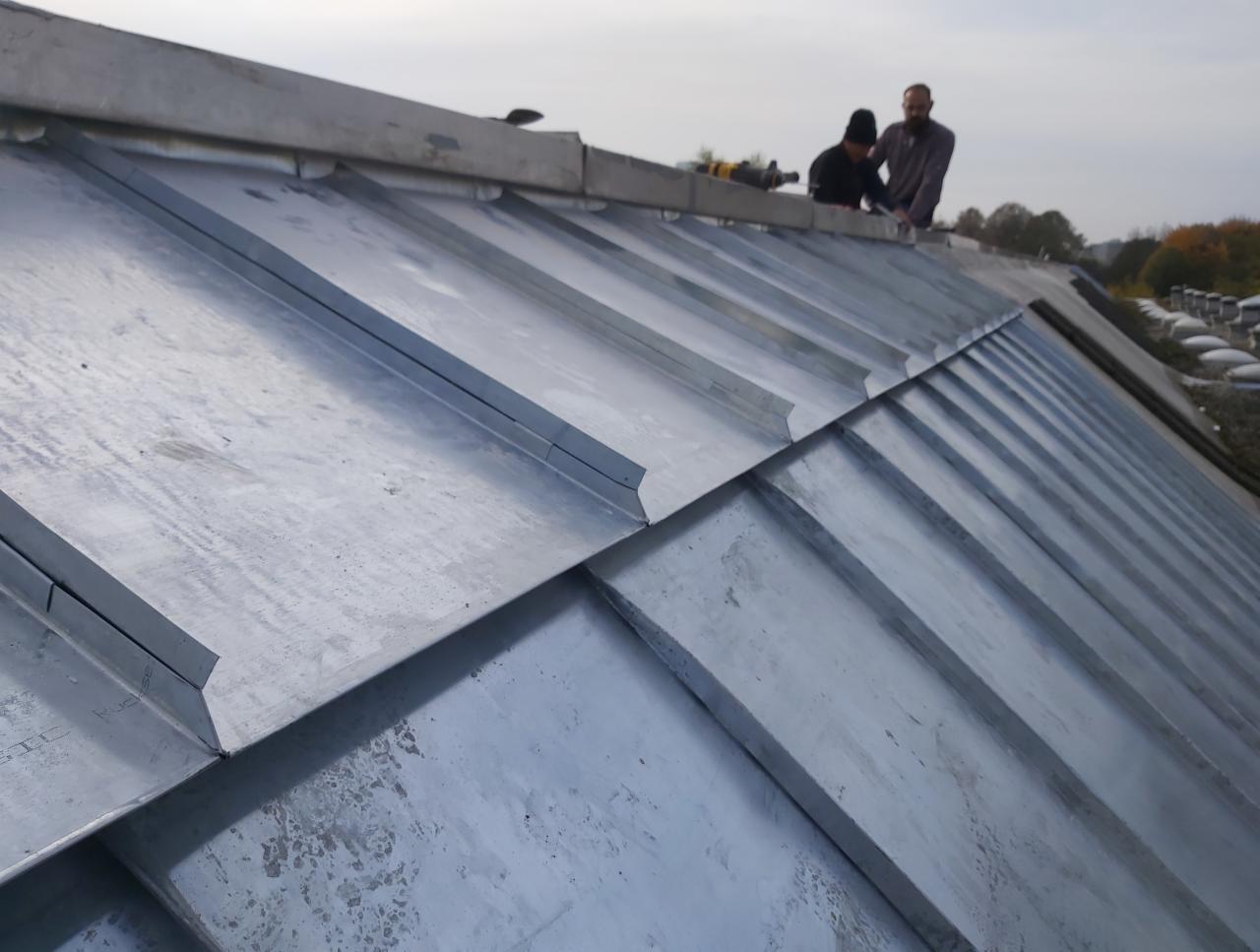 Montaż blachy na dachu z widocznymi dwoma dekarzami i narzędziami, z perspektywy dolnej krawędzi dachu.