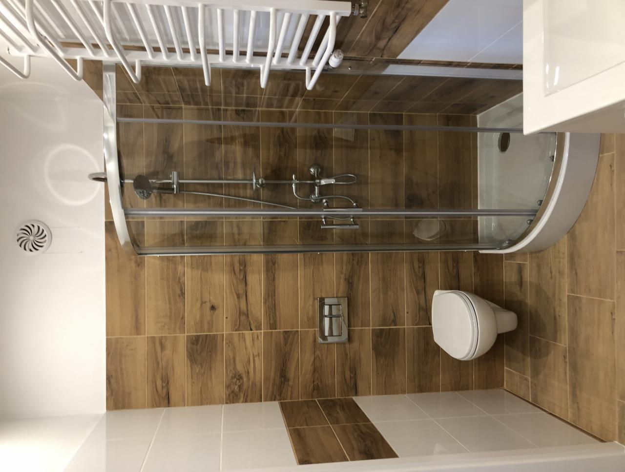 Nowoczesna kabina prysznicowa z drewnopodobnymi płytkami ściennymi i białymi płytkami podłogowymi, widoczny biały grzejnik drabinkowy i toaleta podwieszana.
