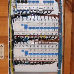 Otwarta, biała skrzynka rozdzielcza z zamontowanymi bezpiecznikami i uporządkowanymi przewodami w różnych kolorach, zamocowana na drewnianej ścianie.