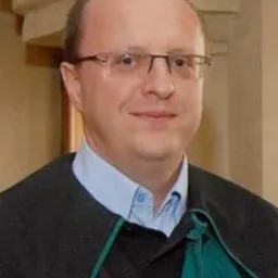 Portret mężczyzny w todze z zielonym wykończeniem, w okularach i jasnej koszuli, na rozmytym tle wnętrza.