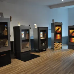 Ekspozycja różnych modeli pieców wolnostojących w salonie, w tym piec z obudową imitującą kamień i piec z miejscem na drewno, widoczne etykiety energetyczne.