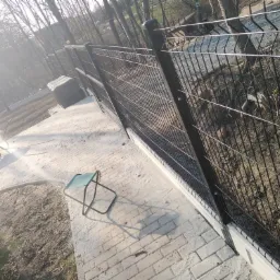 Nowoczesne, czarne ogrodzenie z paneli siatkowych na betonowej podmur&oacute;wce, widziane z perspektywy ogrodu, z w&oacute;zkiem i krzesłem w tle, w słoneczny dzień.