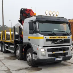 Srebrna ciężarówka z napisem 'POMOC DROGOWA' i dźwigiem na platformie, zaparkowana na placu, obok drewnianych konstrukcji.