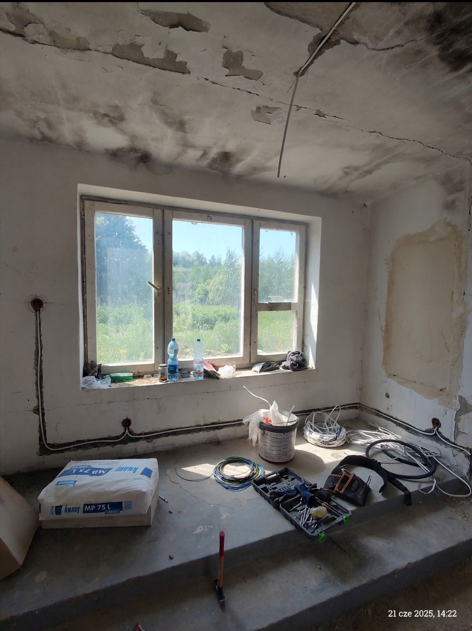 Wnętrze w trakcie remontu: widoczne kable elektryczne w ścianach, narzędzia, worek z zaprawą Knauf MP 75 L. Okno z widokiem na zieleń. Ściany przygotowane do malowania.