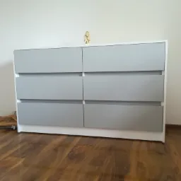 Szara komoda z sześcioma szufladami na drewnianej podłodze, z małą figurką na górze i wieszakami obok.