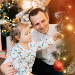 Tata z córeczką dekorują świąteczne drzewko, dziewczynka w bluzie z motywem reniferów dotyka gałązki, w tle ceglana ściana i migoczące światełka