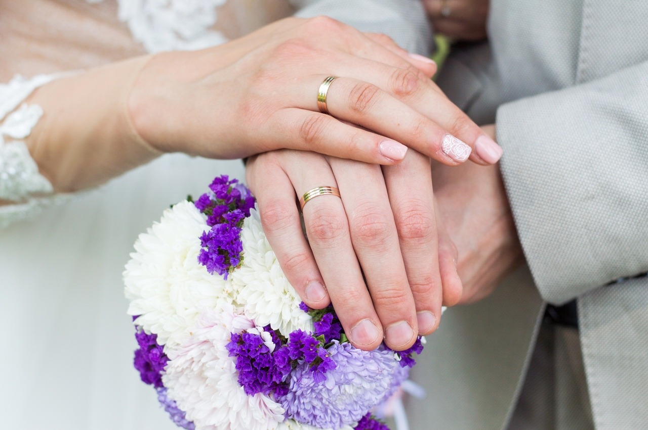 Dłonie pary młodej z złotymi obrączkami spoczywają na bukiecie ślubnym z białych i fioletowych kwiatów; widoczny mankiet szarej marynarki i fragment sukni ślubnej z koronką.