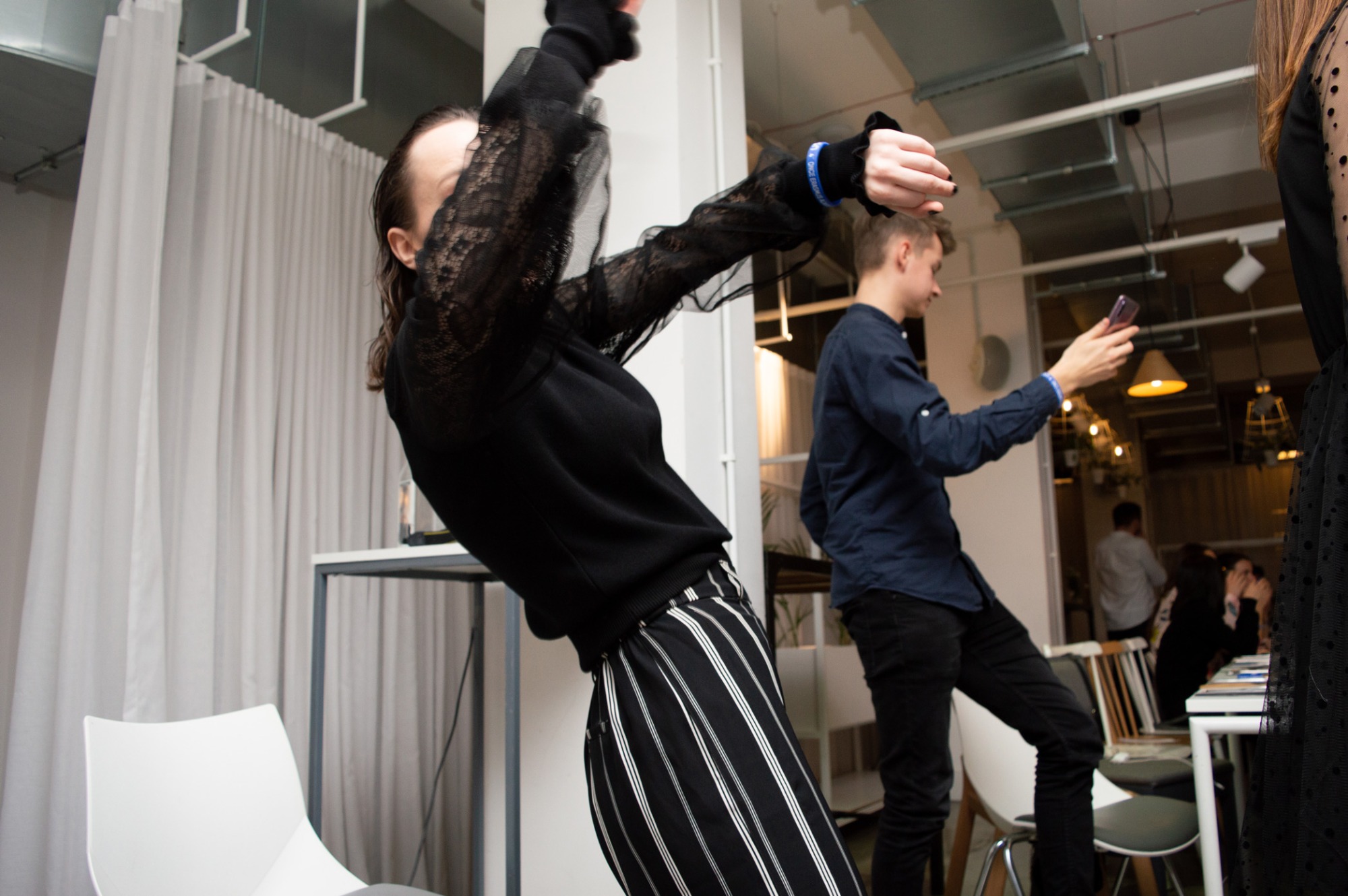 Kobieta w czarnej bluzce z koronkowymi rękawami i spodniach w paski pozuje w dynamicznej pozie na tle jasnego wnętrza z innymi osobami, mężczyzna robi zdjęcie telefonem.