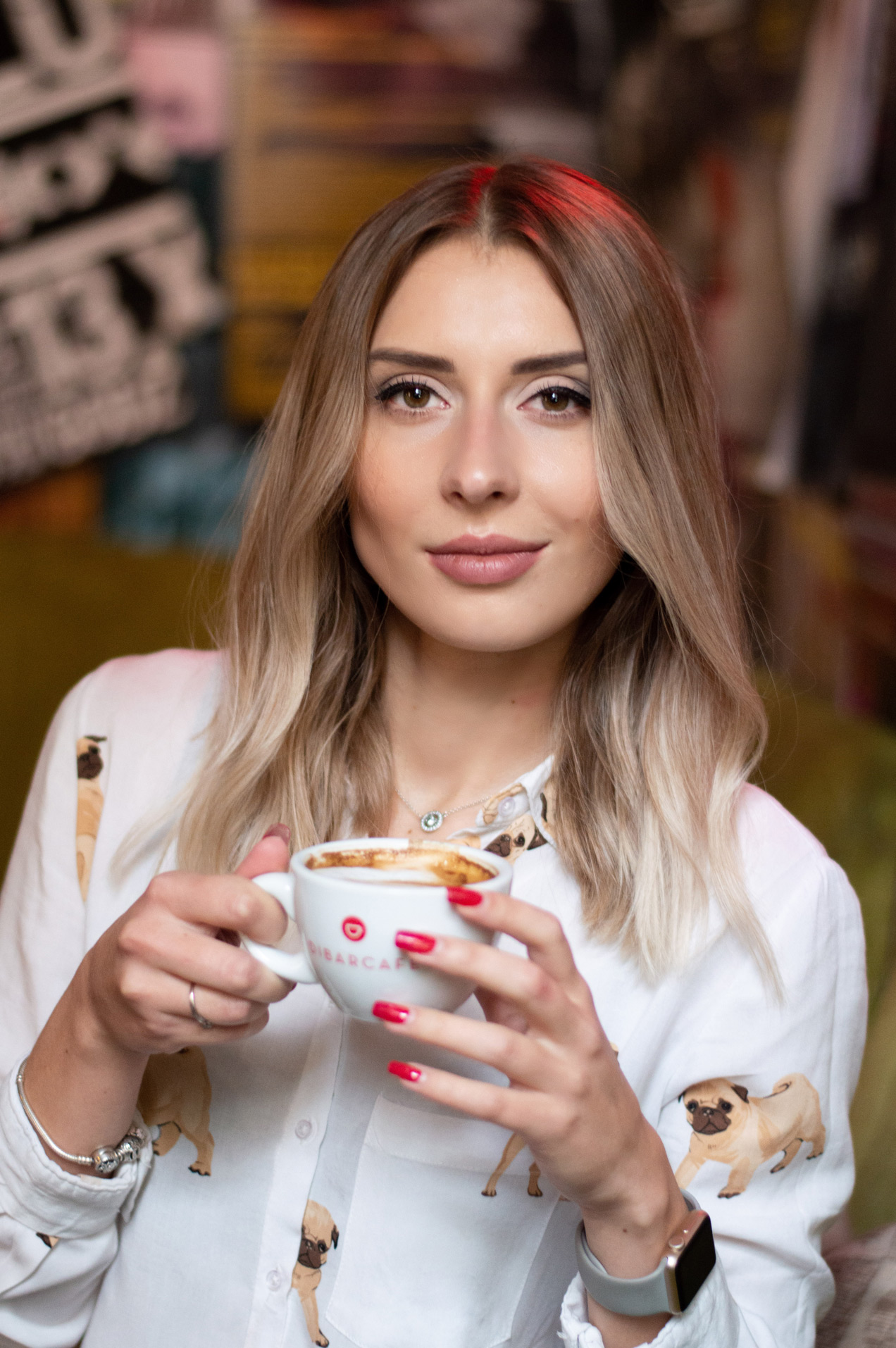 Portret kobiety z blond włosami, trzymającej biały kubek z kawą, ubrana w białą koszulę w mopsy, z czerwonymi paznokciami, na rozmytym tle.