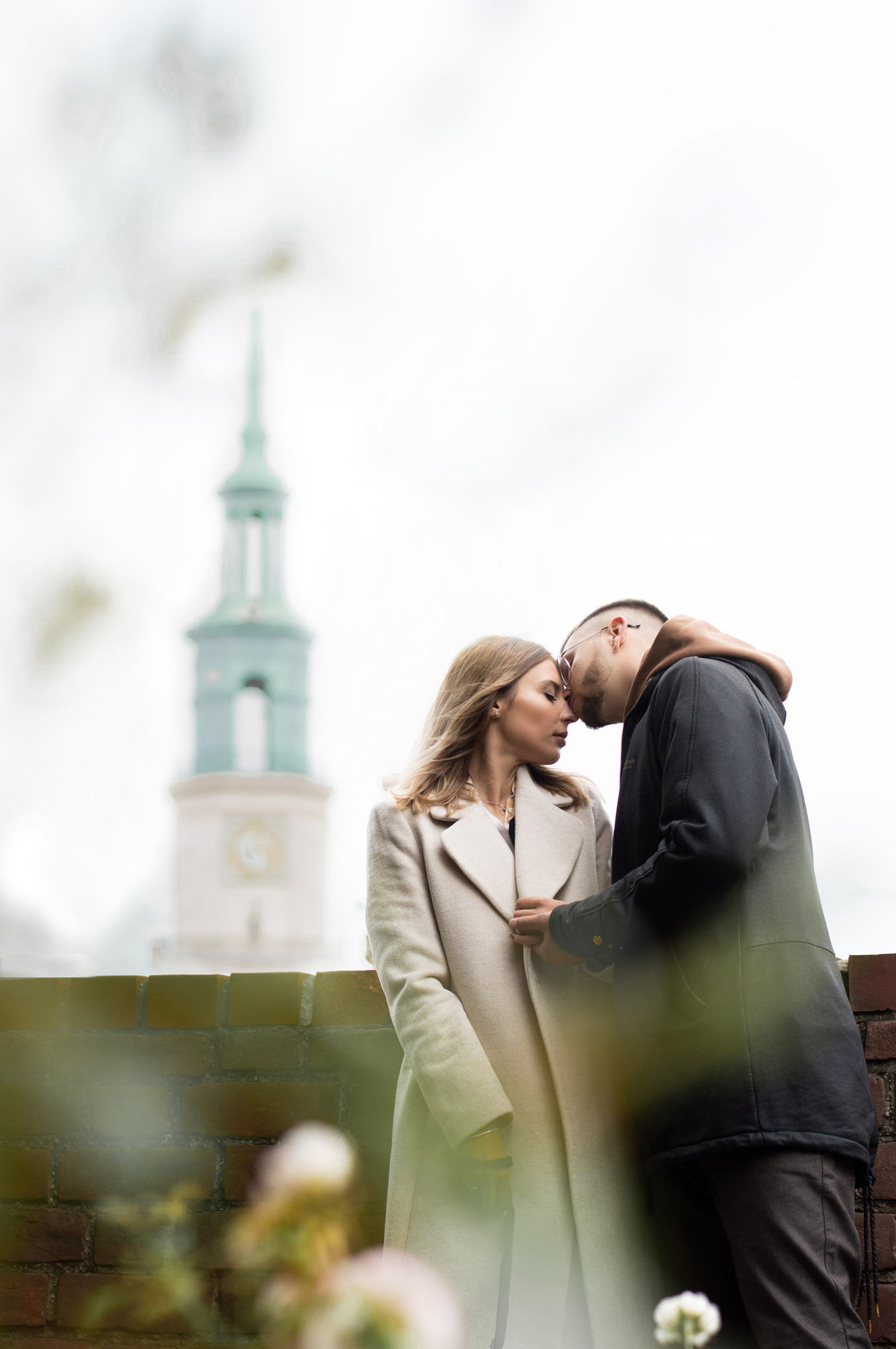Para zakochanych, delikatny dotyk czołami, ona w beżowym płaszczu, on w ciemnej kurtce, ceglana balustrada, w tle wieża kościelna.