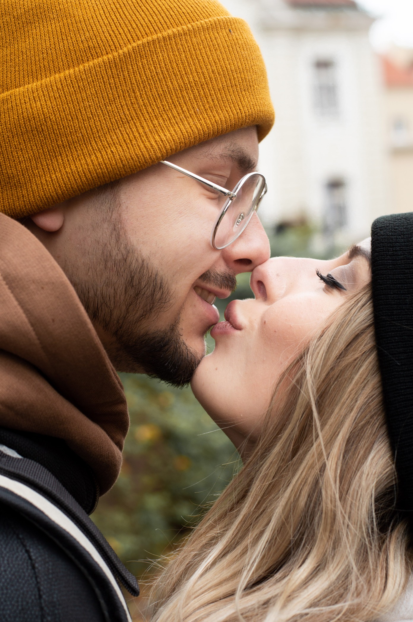 Zbliżenie profilu całującej się pary w czapkach: on w musztardowej, ona w czarnej, oboje z delikatnym uśmiechem, na rozmytym tle miejskim.