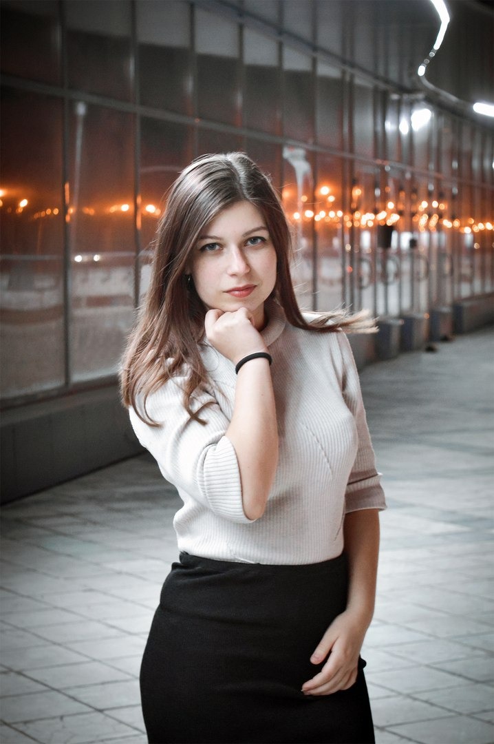 Portret kobiety w jasnym swetrze i czarnej spódnicy, opierającej podbródek na dłoni, na tle przeszklonego korytarza z rozmytymi światłami w tle.