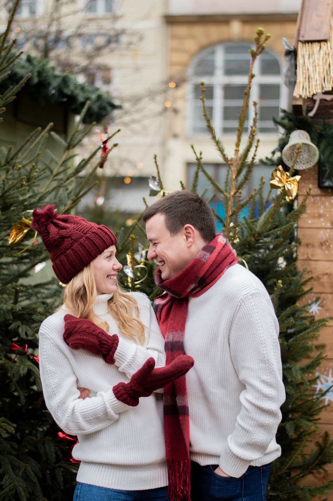 Uśmiechnięta para w białych swetrach i bordowych akcesoriach stoi na tle choinek i świątecznego straganu z dekoracjami.