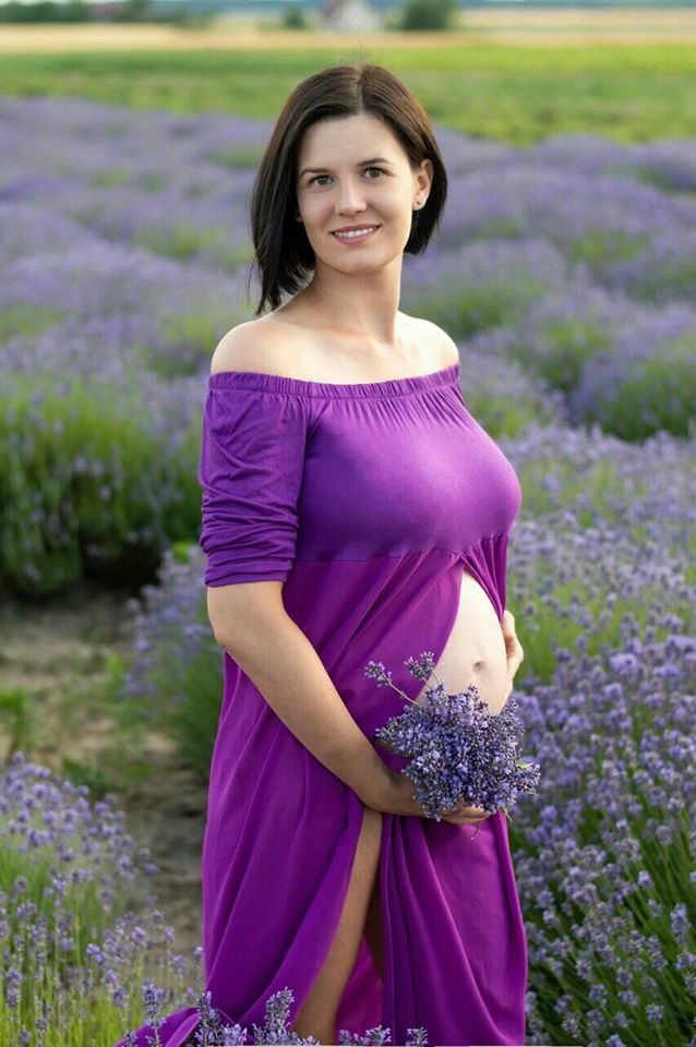 Uśmiechnięta, ciężarna kobieta w fioletowej sukience odsłaniającej ramię, trzyma bukiet lawendy na tle pola lawendy.